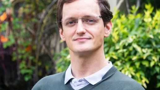 man with glasses, open necked shirt and green jumper, roses bushes behine, looking at the camera