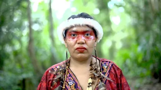 Woman with red face paint across her face, feathered earrings and a red top stands infront of a blurred forest background