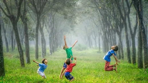 Four children jumping up to catch a football. They are playing on grass inbetween two tall rows of trees