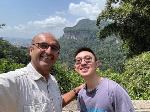 one man with glsses with his hand on the shoulder of another, younger man to this right. They stand in front of jungle and mountains