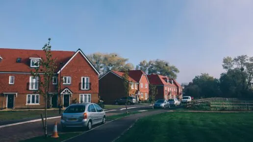 Modern housing alongside a road with parked cars and a grassy area. Mature trees are visible above the roofs of the houses.